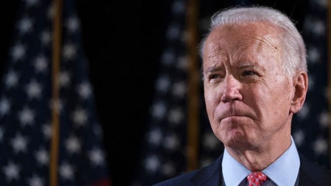 Joe Biden looks into the distance with US flags behind him.