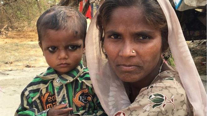 Khazanchi Nath com olhar sério no coloo que é estrelabetsua mãe, que dá um sorriso tímido, com ambiente rústico ao fundo