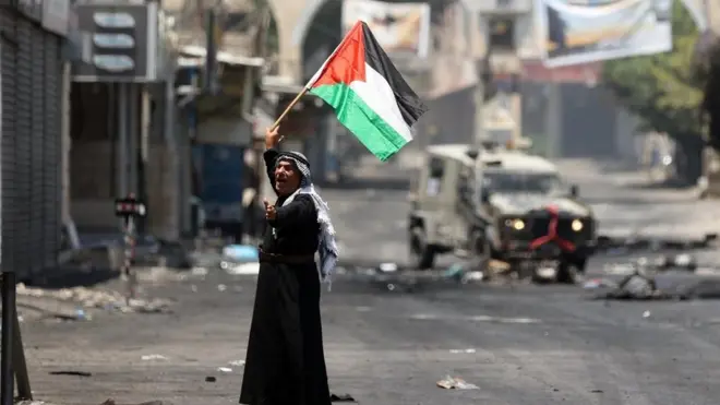 فلسطيني يرفع علم فلسطين أمام عربة عسكرية إسرائيلية أثناء إخراج فلسطينيين من المنطقة