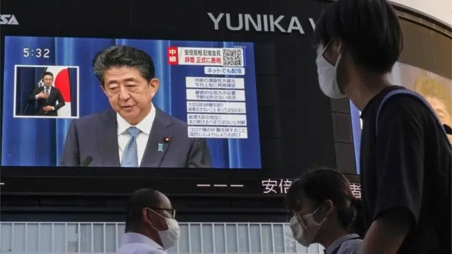 People walk past a display at Shinjuku, showing Japanese Prime Minister Shinzo Abe announcing his resignation during a televised news conference (28 August 2020)