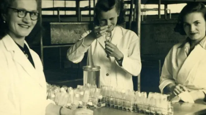Three female scientists in white lab coats smiling in front of some test tubes, one of them is injecting something into a test tube