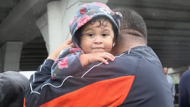 A young girl, looking directly at the camera, is carried by a man through the rain in a flooded neighbourhood