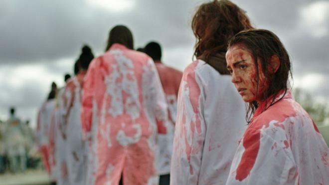 Un fotograma de la película que muestra a la protagonista con la cara llena de sangre y una bata blanca salpicada también de sangre haciendo una fila detrás de otras personas.