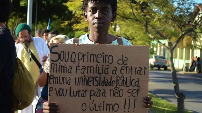 Jovem negro segurando um cartaz que diz 'Sou o primeiro da minha família a entrar1xbetmnuma universidade pública e vou lutar para não ser o último'