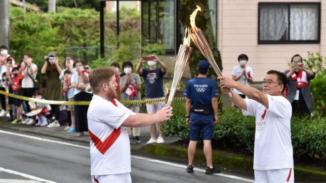 Torchbearers, Australian Shaun Ballinger (L) and Japanese Atsushi Kawaguchi (R) hand over the flame of the Olympic torch during the Tokyo 2020 Olympic Games Torch Relay in Gotemba