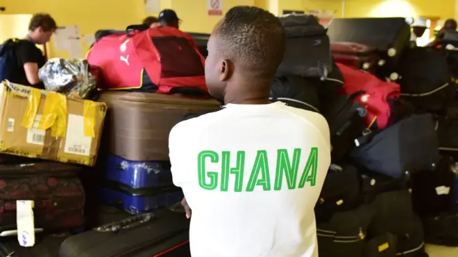 A man wearing a tee shirt on which is written 'Ghana' on the back, stands next to the pile of suitcases belonging to Ghana's national football team players and officials, at the airport