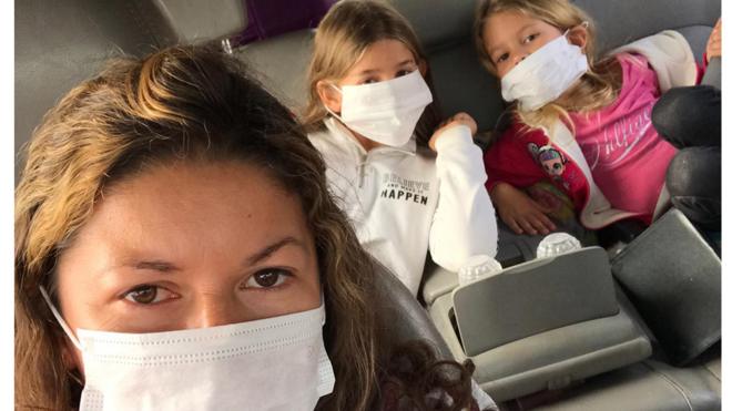 Maria, com máscara, tira selfie no carro com as duas filhas pequenas no bancolei casa de apostastrás do carro, também com máscaras