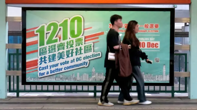 香港元朗某港鐵輕鐵站一對男女走過一幅政府宣傳區議會選舉的海報（BBC中文圖片19/11/2023）