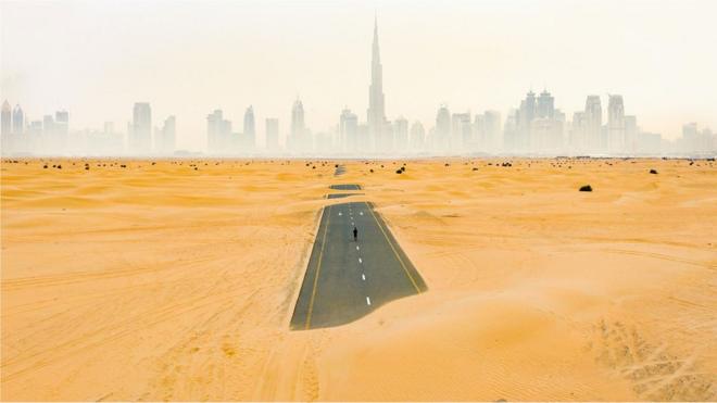 Uma pessoa caminha por uma estrada deserta no desertoroleta da sorte googleDubai cobertaroleta da sorte googledunas,roleta da sorte googledireção a uma paisagem urbana