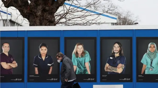 ชายคนหนึ่งเดินผ่านป้ายขนาดใหญ่หน้าโรงพยาบาลแห่งหนึ่งแสดงให้เห็นภาพบุคลากรต่าง ๆ ของ NHS