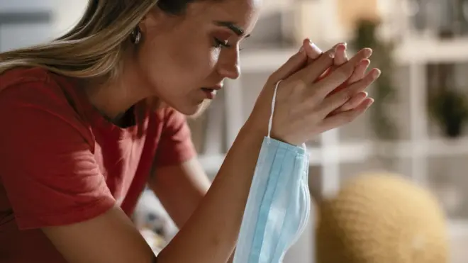Una mujer preocupada con una mascarilla en la mano.
