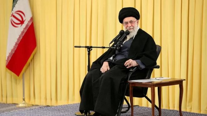 Али Хаменеи на фоге желтого занавеса, радом с иранским флагом