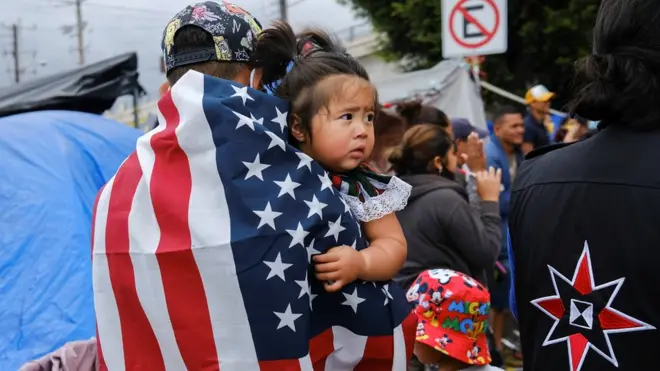 мигранты из гондураса Honduran migrant Kevin, wearing a U.S. flag, holds his daughter Keiry, during a multicultural activity at a makeshift camp at the El Chaparral border port of entry with the U.S., in Tijuana, Mexico April 22, 2021. Picture taken April 22, 2021. REUTERS/Toya Sarno Jordan