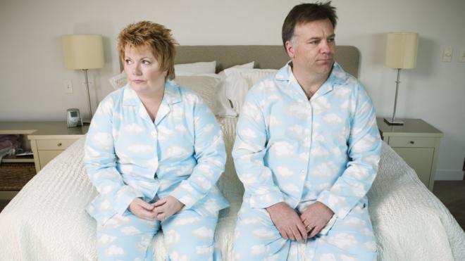 Hombre y mujer vestidos iguales y sentados en el borde de la cama con cara de descontento.