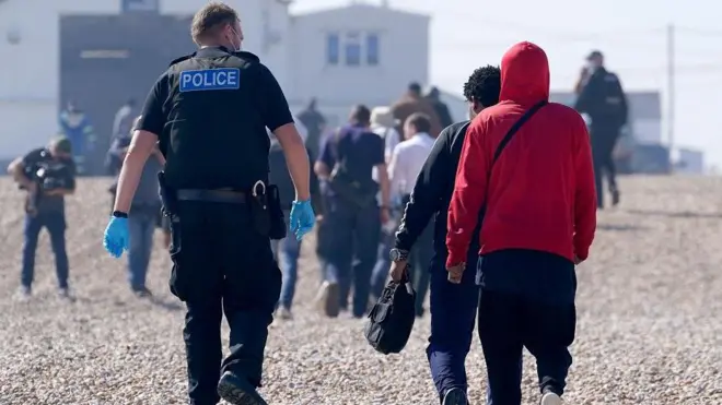 Полицейский сопровождает группу людей, предположительно мигрантов, на берег со спасательной шлюпки в Дунгенессе, графство Кент. Все люди на фотографии, включая полицейских, находятся к камере спиной.
