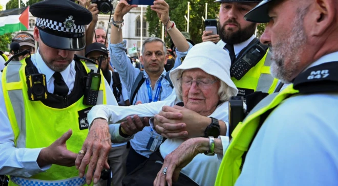 Gözaltına alınan şapkalı ve yaşlı kadının yanında üç polis var. Kadın gözlerini kapatmış.