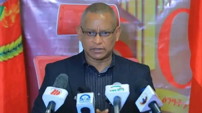 ኣብ ቅድሚኡ ማይክሮፎናት ሚዲያ ዝተተኽለሉ ደብረጽዮን ገብረሚካኤል መግለጺ እናሃበ፡ ኣብ ድሕረባይታኡ ባንዴራታትን ኣርማታትን ህወሓት ይርአ