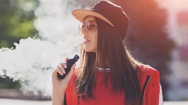A young woman, wearing a cap and sunglasses and dressed in a red top, exhales steam from a vape. She is standing outdoors in the sunshine. 