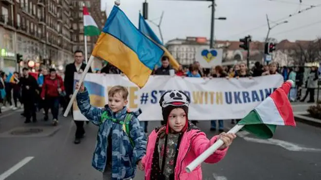 Двоє дітей йдуть попереду демонстрантів по вулиці у Будапешті, тримаючи у руках прапори України на Угорщини. Позаду них - люди з гаслами на підтримку України.