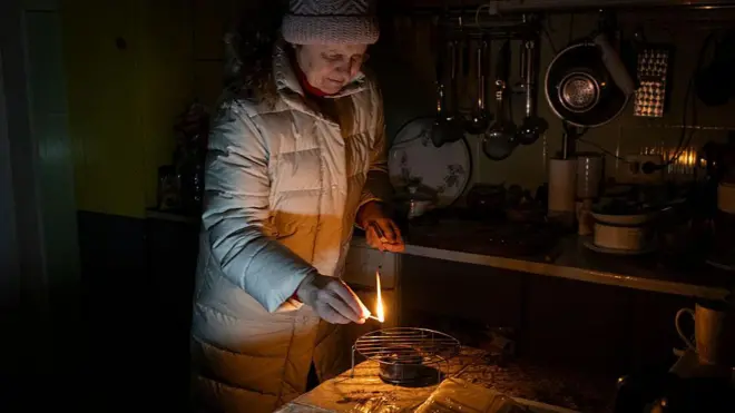 Жінка у світлій зимовій куртці та шапці запалює "окопну" свічку під металевою решіткою, що стоїть на столі в темній кухні. На задньому плані видно підвішене кухонне приладдя, тарілки та посуд, а також стільницю з різними предметами. Освітлення слабке, джерелом світла є сірник.