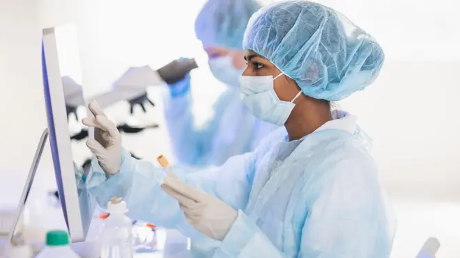 A masked female researcher looking at a computer screen in a lab environment
