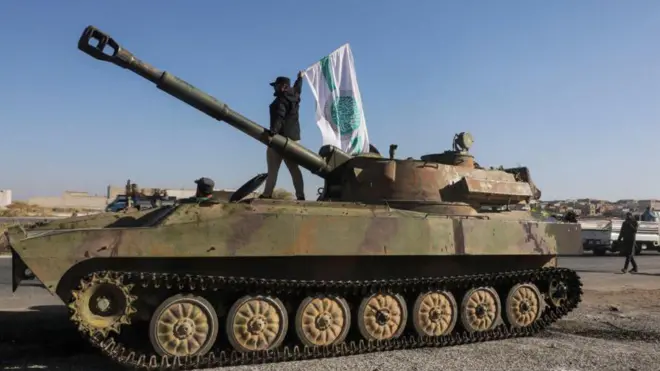 أحد أفراد المعارضة المسلحة يلوح بعلم هيئة تحرير الشام فوق دبابة في سراقب، محافظة إدلب، الأول من ديسمبر/كانون الأول 2024