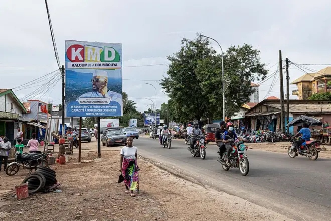 Des motos et véhicules sur une route avec à côté un panneau géant sur lequel est mis une affiche du chef de la junte en Guinée.