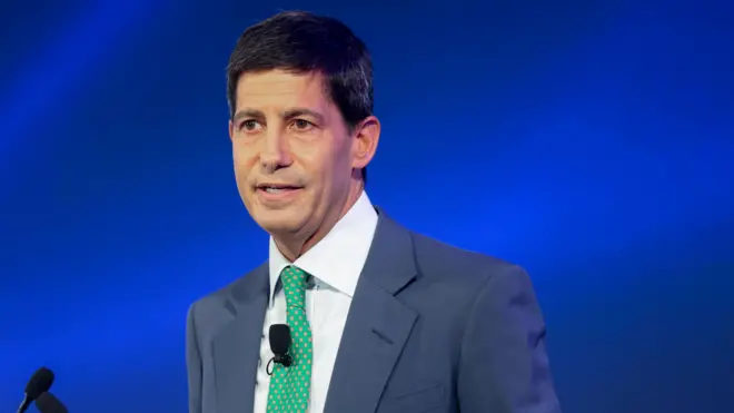 Кевин Уорш, мужчина средних лет с темными короткими волосами и карими глазами, без бороды, в пиджаке с зеленым галстуком.