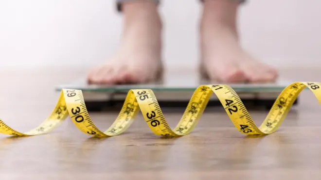 Imagem desfocada dos pés de uma pessoa se pesando em uma balança ao fundo, com uma fita métrica em primeiro plano