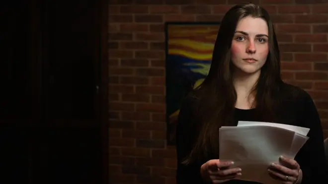 Michele olhando sério para cama, em sala de casa, segurando papelada