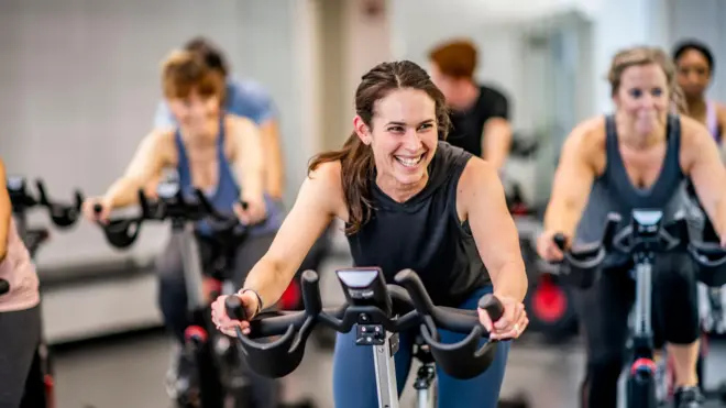 -Mulher sorrindo faz exercício em uma bicicleta ergométrica, enquanto outras pessoas aparecem fora de foco fazendo o mesmo exercício