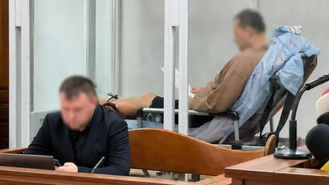 Фото із зали суду - на передньому плані сидить чоловік, на задньому - інший чоловік напівлежить на медичній кушетці, їхні обличчя розмиті