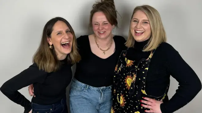 Three women are standing together laughing. The woman on the left haas dark hair and is wearing a black top, the one in the middle has dark hair and is wearing a black top and blue jeans. The woman on the right has blonde hair and is wearing a black top underneath overalls with pictures of the sun and moon on them