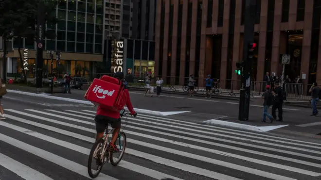 Um entregador anda de bicicleta enquanto faz uma entrega na Avenida Paulista, em São Paulo. Ele atravessa a faixa de pedestre e tem uma mochila vermelha com a marca iFood 