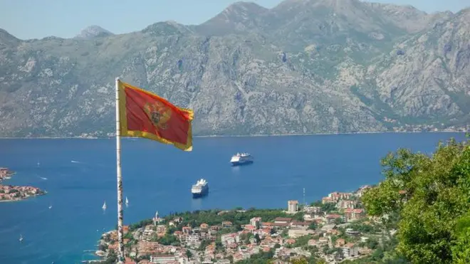 Yüksek bir yerden çekilen fotoğraf Karadağ bayrağı arkasında deniz manzarası ve dağları gösteriyor. 