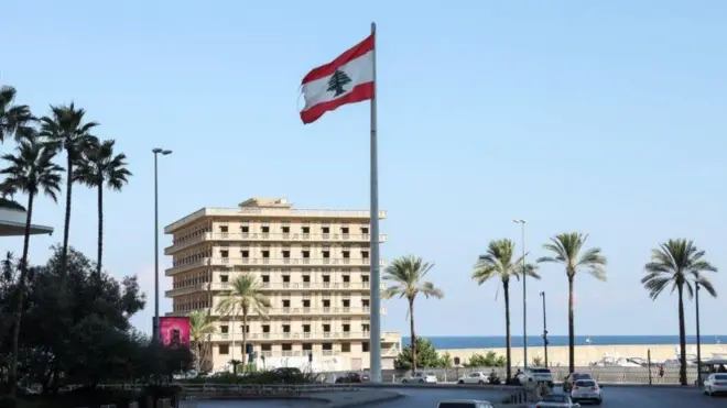 مركبات تسير بالقرب من العلم اللبناني الذي يرفرف في بيروت قرب الشاطئ، الصورة التقطت نهاراً.