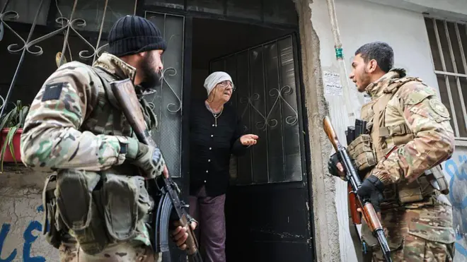 İki askeri kamuflaj kıyafetleriyle bir evin kapısının önünde duruyor, açık kapıdan dışarı doğru ileri yaşlı bir kadın, askerlerden sağdakiyle konuşuyor.