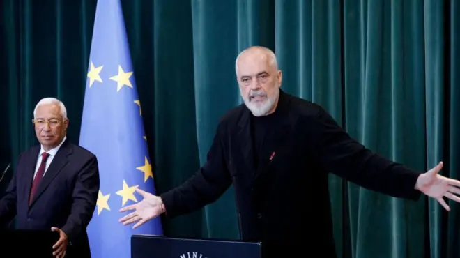еди рама у црној мајици и панталонама, иза њега са леве стране седи мушкарац у оделу и кравати