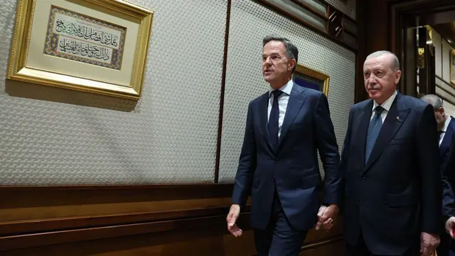 Erdoğan ve Rutte el ele duvarda Arapça yazıların olduğu bir koridorda yürüyor.