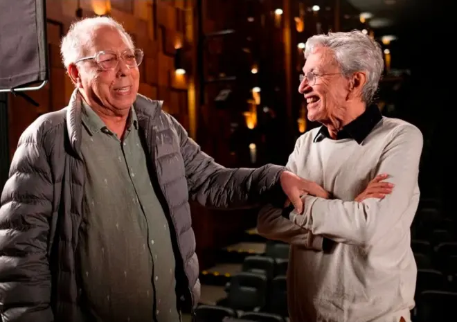 Os cantores Jards Macalé e Caetano Veloso dando risada, enquanto Macalé toca o braço de Caetano afetuosamente