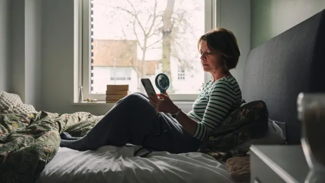 Kobieta w średnim wieku siedzi na łóżku. W dłoniach trzyma telefon komórkowy i przenośny wentylator. Przez okno za nią widać czerwone dachy domów i drzewa. 