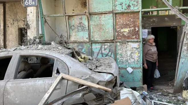 Літня жінка стоїть у під'їзді пошкодженого житлового будинку. Ліворуч від неї стоїть сильно пошкоджений автомобіль без вікон. Навколо розкидані уламки.