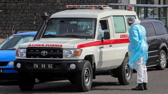 bir ambulansın yanında sırtı dönük bir halde ve koruyucu giysiyle duran bir sağlık görevlis