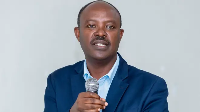 Philibert Gakwenzire, Perezida wa IBUKA mu Rwanda, afashe indangururamajwi mu kiganza arimo kuyivugiramo. Yambaye ikositimu y'ubururu n'ishati y'ubururu bwerurutse. 
