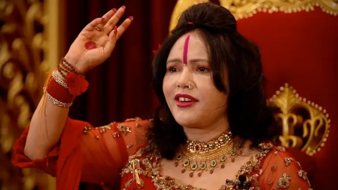 Radhe Maa with dark hair, dressed in red and gold with jewelry, sitting in a ornate chair and pointing upwards with her right hand