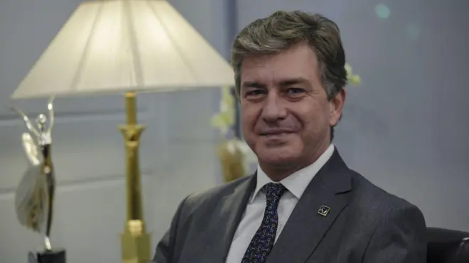 Gülümseyen bir adam, kameraya bakıyor. Arka planda ödül ve bir lamba var