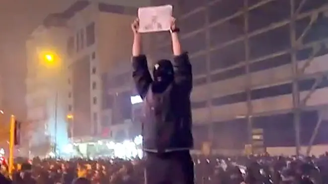 A masked protester stands on a platform above crowds at night in Kaj Square, in north-west Tehran. on 9 January 2026. They are holding a black and white photo above his head of the last shah's son, Reza Pahlavi. Light from buildings, streetlamps and shop fronts are illuminating the area. The protester's mask is black with a white mark on the front, with three holes cut out for their eyes, mouse and nose.