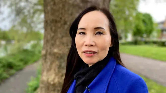 A woman in a blue coat standing in front of a tree in a park looking at the camera.
