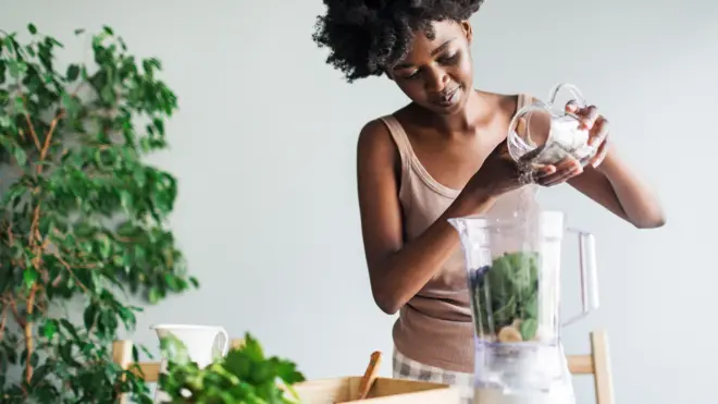 Stock photo shows a woman adding chia seends into a smoothie at home, there is a plant in the background.