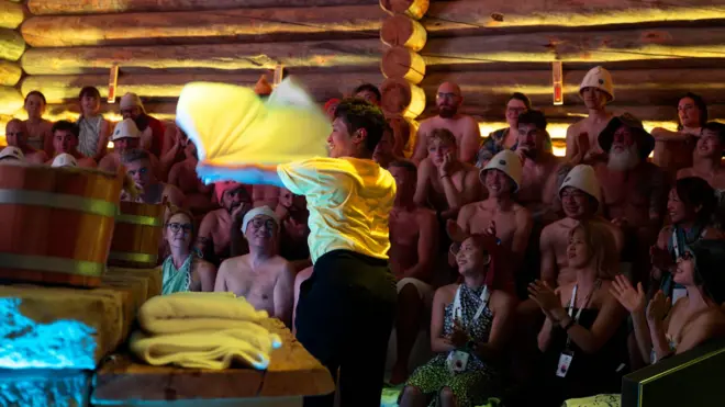 Ludzie siedzą w drewnianej saunie, przyglądając się mężczyźnie, który macha białym ręcznikiem.